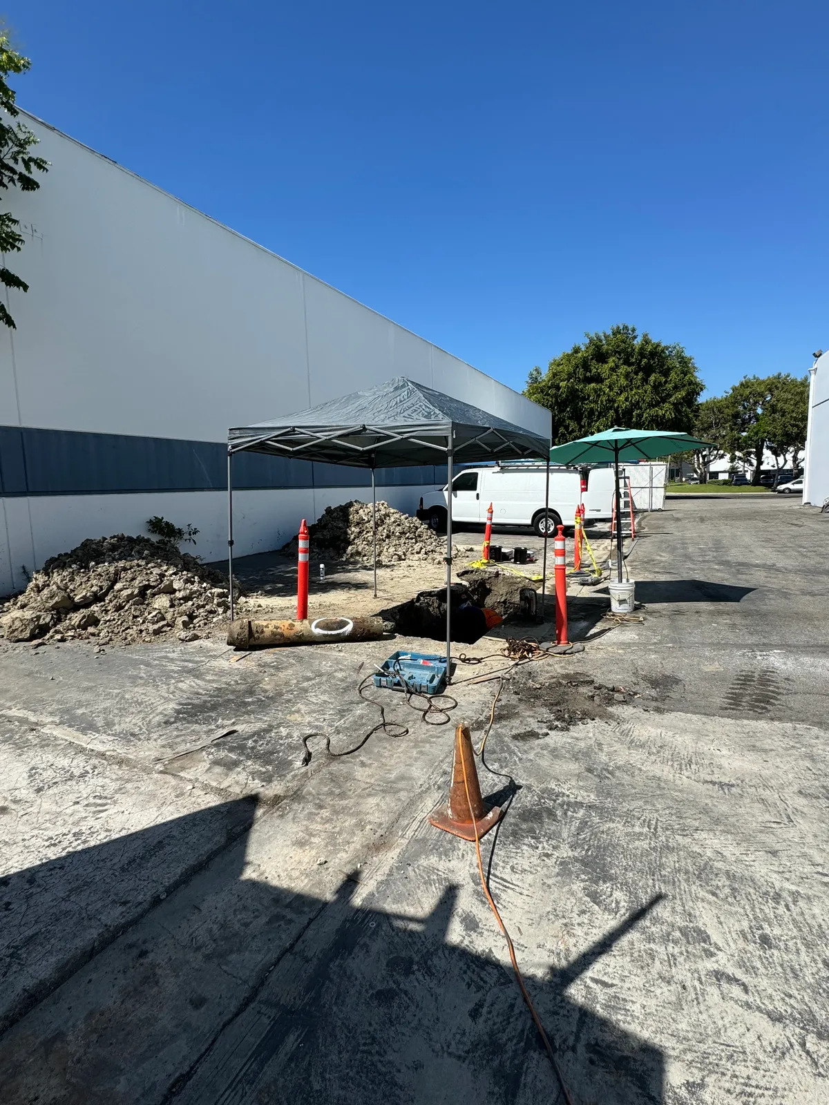 Excavated sewer repair under a canopy in a commercial parking lot with dirt piles and traffic cones